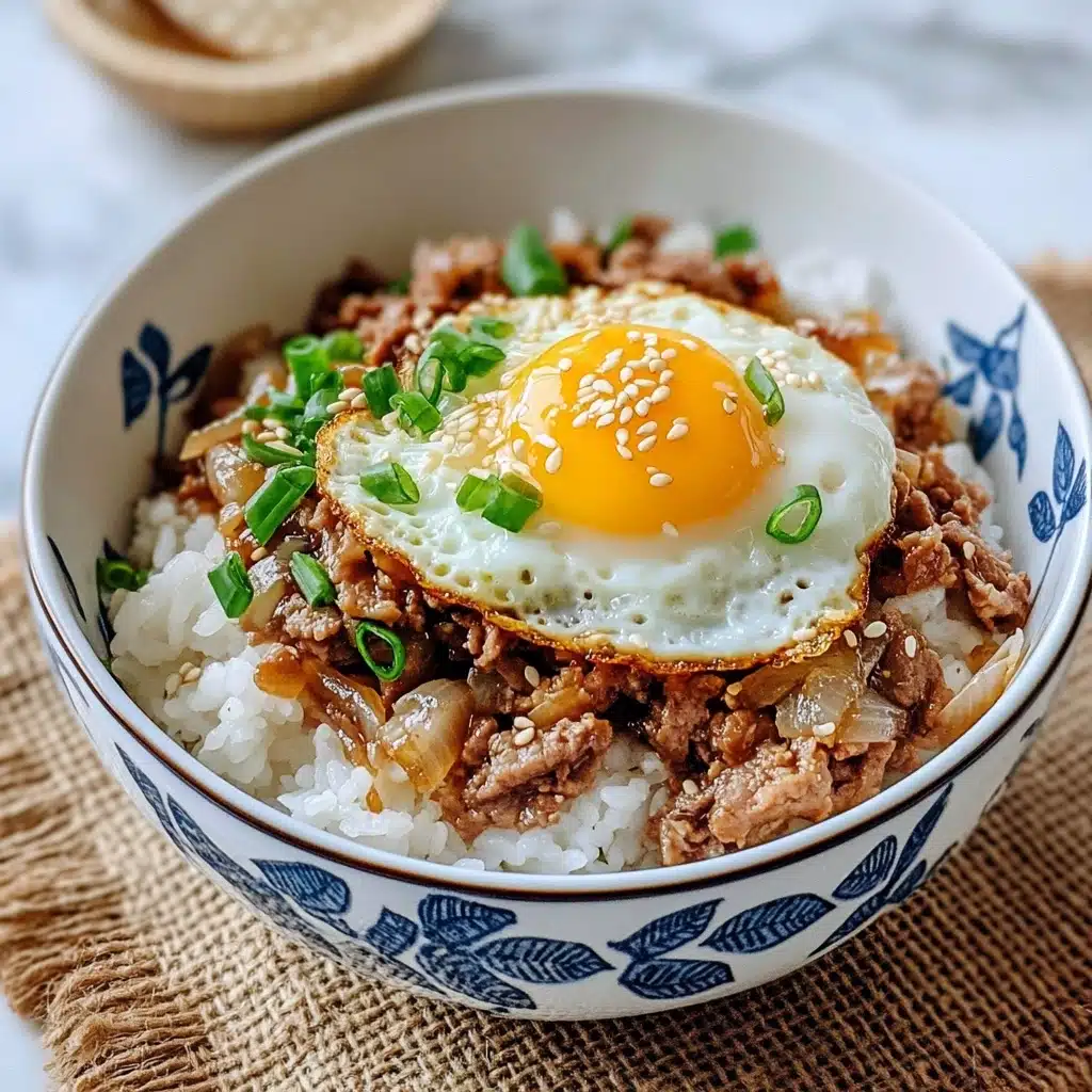 Japanese Beef & Rice Bowls Gyudon, gyudon recipe, Japanese beef bowl, quick Japanese dinner, savory beef rice bowl A white bowl with blue leaf patterns holds a three-layer dish. The bottom layer is white cooked rice with a soft texture. Above the rice is a layer of cooked brown meat mixed with translucent cooked onions, garnished with small green sliced scallions and small white sesame seeds. On top of this is a bright yellow sunny-side-up fried egg with a shiny yolk and lightly crisp white edges, sprinkled with more sesame seeds and some scallion slices. The bowl sits on a natural brown burlap cloth with a white marbled background.