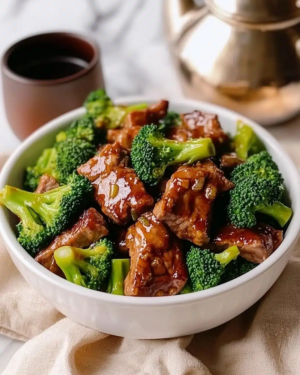 Beef and Broccoli Stir-Fry, Chinese Beef Stir-Fry, Easy Asian Beef Recipe, Quick Weeknight Dinner, Healthy Beef and Broccoli A white bowl is filled with two main layers: bright green broccoli florets and slightly glossy brown beef slices mixed together, with broccoli pieces evenly spread over and under the beef on all sides. The bowl sits on a soft beige cloth, placed on a white marbled surface. To the upper left of the bowl, there is a shiny metal teapot, and to the upper right, a cup along with two wrapped fortune cookies and a pair of dark brown chopsticks lying next to a fork. The light is soft and natural, highlighting the fresh texture of the broccoli and the cooked shine on the beef.