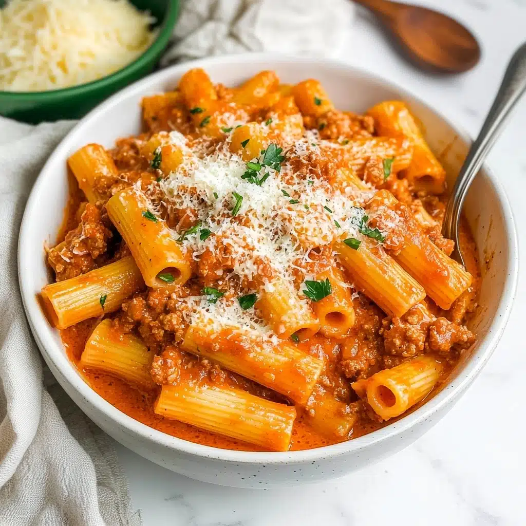 Date Me Creamy Sausage Rigatoni, creamy sausage pasta, easy Italian pasta recipe, comfort food dinner, quick one-pot pasta A white bowl filled with creamy orange-red sauce pasta, showing rigatoni pasta tubes covered in a thick sauce mixed with browned ground meat pieces. On top, a light sprinkle of grated white cheese and small green herb leaves adds color contrast. A spoon with a long silver handle is partially inside the bowl on the right side. Behind the bowl, there is a small green dish holding more grated cheese and a wooden spoon with dark brown grain resting next to it. The bowl and items are placed on a white marbled surface with a pale cloth on the left side.