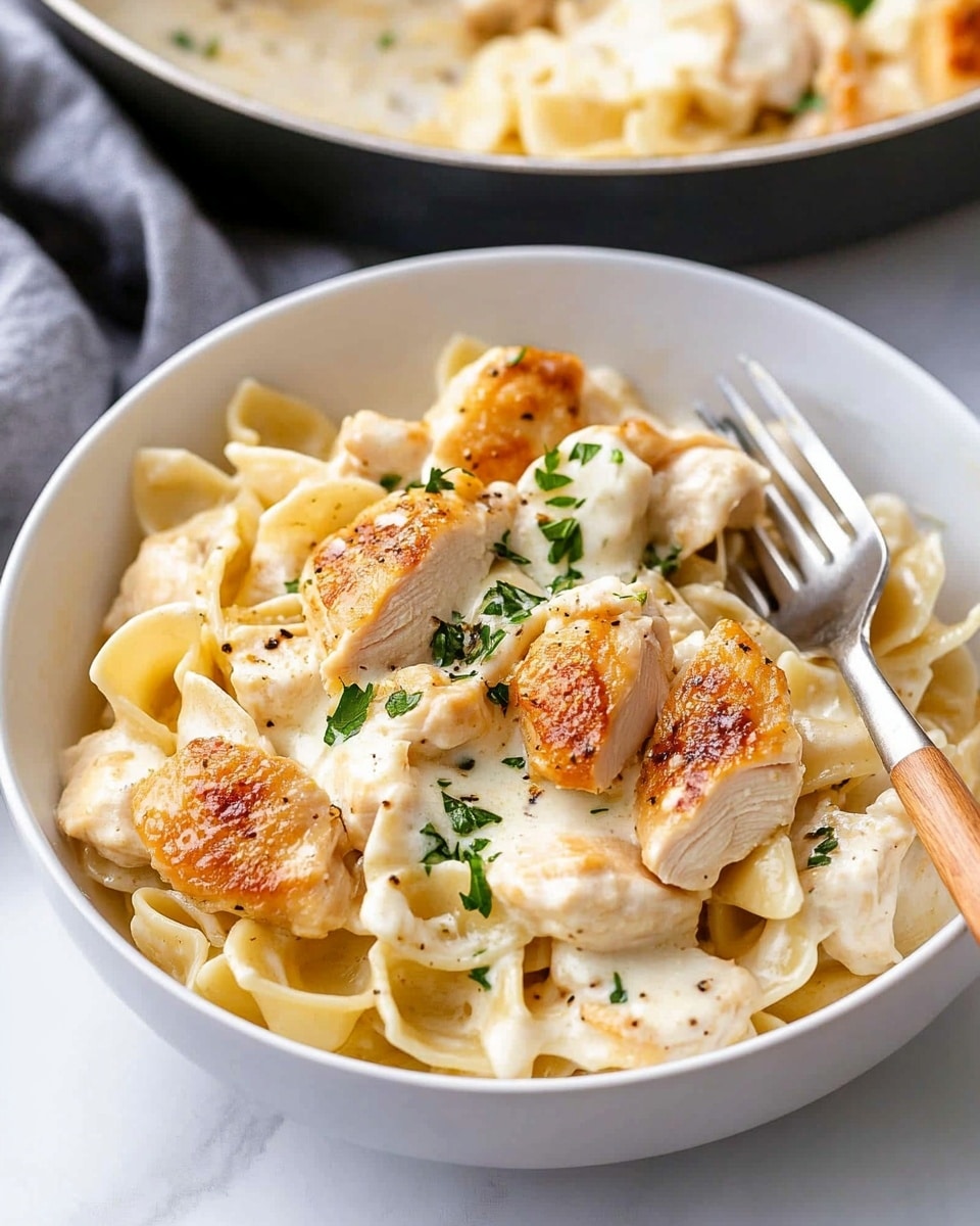 Chicken Tortellini Alfredo, creamy chicken tortellini, quick Alfredo pasta, cheesy chicken tortellini dinner, easy Italian Alfredo recipe The image shows a close-up of a dish with about two layers: the first layer is soft, folded pasta with a creamy white sauce that looks smooth and thick, creating a rich base. On top, scattered unevenly, are thick pieces of cooked chicken, golden-brown on the outside with a juicy, white inside. Small green bits of parsley are spread across the chicken and pasta, adding a fresh pop of color. The whole dish sits on a white plate over a white marbled surface, giving a clean and bright look.