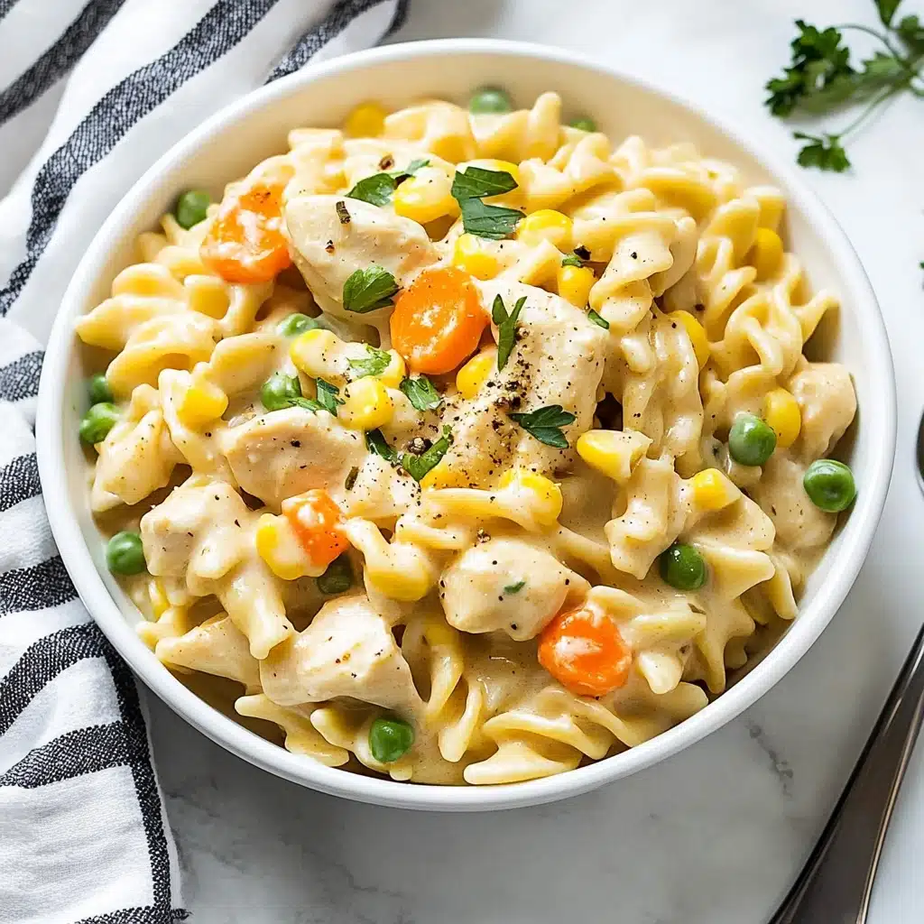 Instant Pot Cheesy Chicken, Noodles, and Vegetables, cheesy chicken pasta recipe, quick one-pot chicken dinner, hearty Instant Pot chicken dish, easy cheesy chicken and noodle recipe A white bowl is filled with creamy pasta made of pale yellow noodles mixed with chunks of light beige chicken, bright orange carrot pieces, yellow corn kernels, green peas, and green beans, all coated in a smooth sauce. Small green herb leaves are scattered on top, and a few black pepper specks are visible. The bowl rests on a white marbled surface next to a white and black striped cloth, with a shiny silver spoon placed beside it.