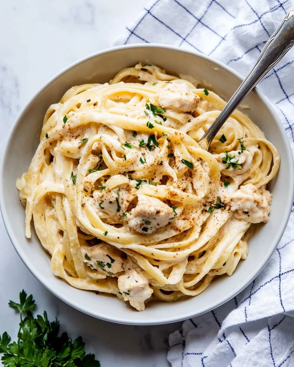 One Pot Cajun Chicken Alfredo Pasta, Cajun chicken pasta, creamy Cajun pasta, easy one pot pasta, quick weeknight dinner The image shows a close-up of a white pot filled with creamy fettuccine pasta mixed with small pieces of chicken. The pasta is light yellow, coated in a smooth light cream sauce, and scattered with finely chopped green herbs. The chicken pieces are white with a slight golden brown edge and sprinkled with a light dusting of paprika or similar seasoning. A wooden spoon is partly visible stirring the dish against a white marbled background.