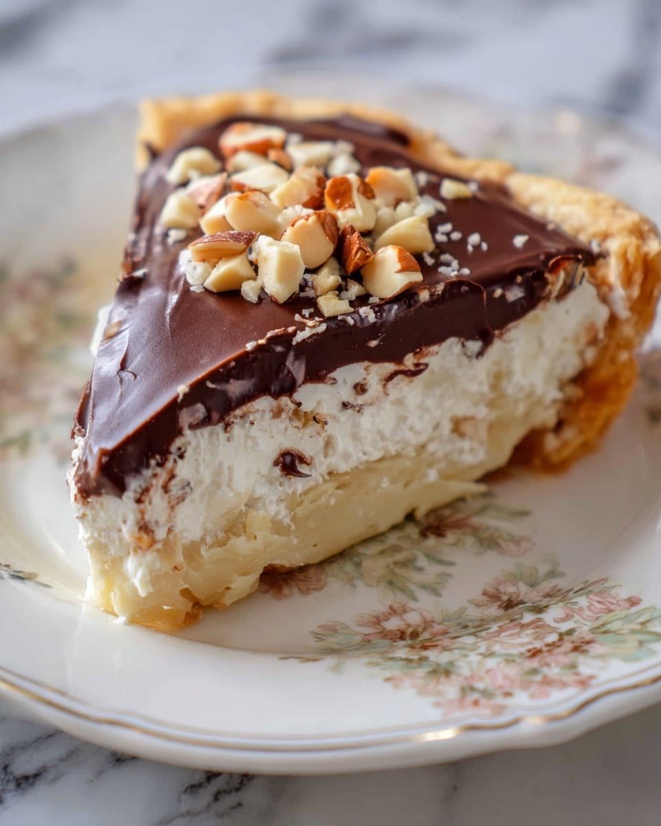 A slice of pie on a white plate with a floral pattern. The pie has three visible layers: the bottom layer is a pale, flaky crust that looks soft and crumbly; the middle layer is thick and creamy, white with some specks inside; the top layer is smooth, dark chocolate with a glossy finish and a small pile of chopped nuts on one corner. The edges of the crust are slightly raised and golden. The photo is taken on a white marbled surface. Photo taken with an iphone --ar 4:5 --v 7