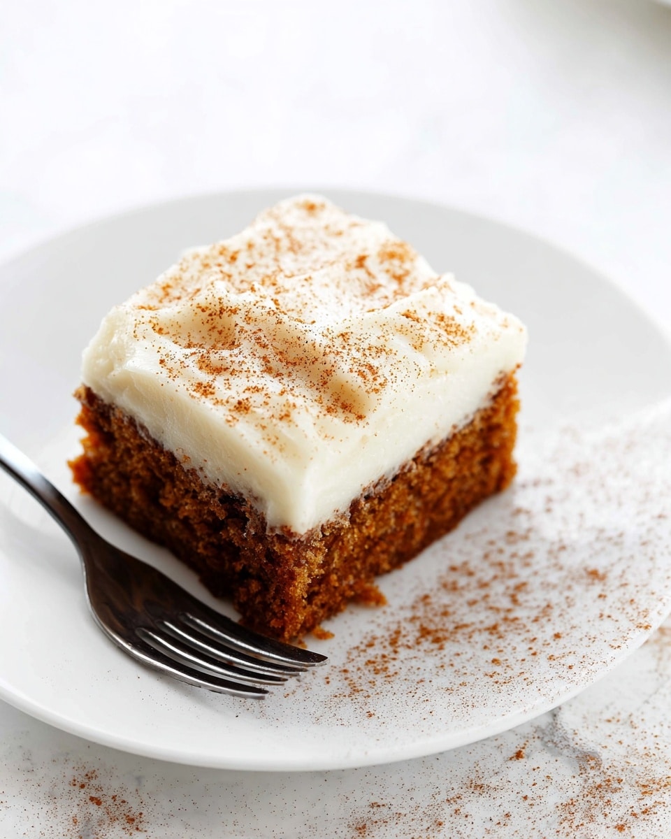 A square piece of moist brown cake sits on a white plate with a silver fork beside it, showing a slightly crumbly texture. The cake has one thick layer of creamy white frosting on top, sprinkled lightly with fine brown spice, likely cinnamon, that is also dusted around the plate. The background and surface beneath the plate are a smooth white marbled texture. Photo taken with an iphone --ar 4:5 --v 7