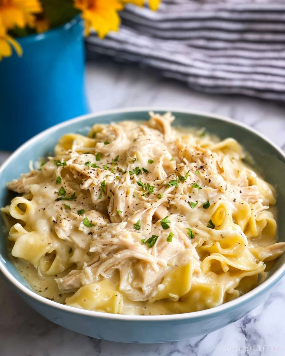 The image shows a white bowl filled with creamy chicken and noodles. There are several layers visible: at the bottom, soft noodles covered by a thick creamy sauce that is pale yellow. On top of that, shredded pieces of light brown chicken are mixed with more noodles and sauce. The sauce looks smooth and glossy, with some specks of black pepper and small bits of green herbs scattered over the dish. The edges of the bowl frame the food tightly. The background is a white marbled surface with a blurred striped cloth and a hint of yellow flowers in a blue pot. Photo taken with an iphone --ar 4:5 --v 7