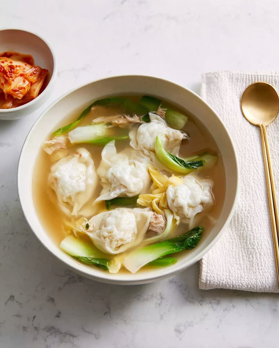 A white bowl filled with clear broth soup contains five white dumplings with slightly wrinkled texture floating on top. There are light yellow egg ribbons and green vegetable stalks scattered around the dumplings, along with small pieces of light brown meat. The bowl sits on a white marbled surface, with a white bowl of reddish kimchi nearby. On the right, a white cloth napkin holds a gold spoon and pair of gold chopsticks. Photo taken with an iphone --ar 4:5 --v 7