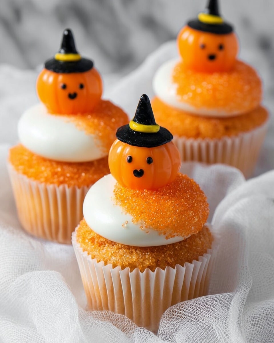 Three orange cupcakes with white paper wrappers sit on a soft white gauze cloth over a white marbled surface. Each cupcake has two main layers: the base is orange cake, and on top is a swirl of white frosting. Part of the frosting is coated with bright orange sugar crystals on one side. Each cupcake is decorated with a small orange pumpkin made of icing or fondant, featuring carved smiling faces in yellow and wearing small black witch hats. Photo taken with an iphone --ar 4:5 --v 7