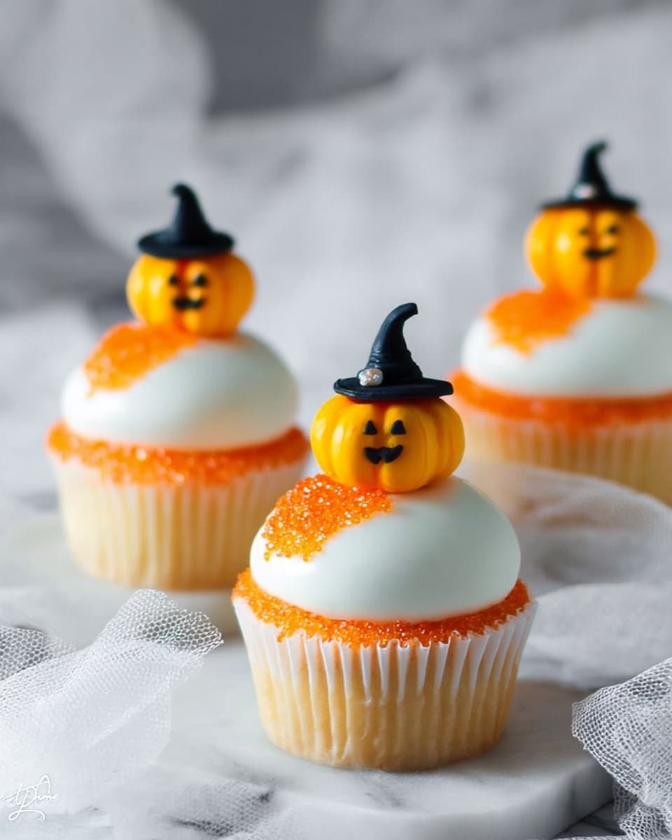 The image shows three cupcakes on a white marbled surface with a white gauzy cloth underneath. Each cupcake has a white base liner and is topped with a smooth white frosting layer. On one side of the frosting, there is a sparkling orange sugar decoration that looks like small crystals. Sitting on top of each cupcake is a small pumpkin decoration with a carved face in yellow and a tiny black witch hat, giving a Halloween theme. The cupcakes are arranged with two in the back and one closer in the front. Photo taken with an iphone --ar 4:5 --v 7