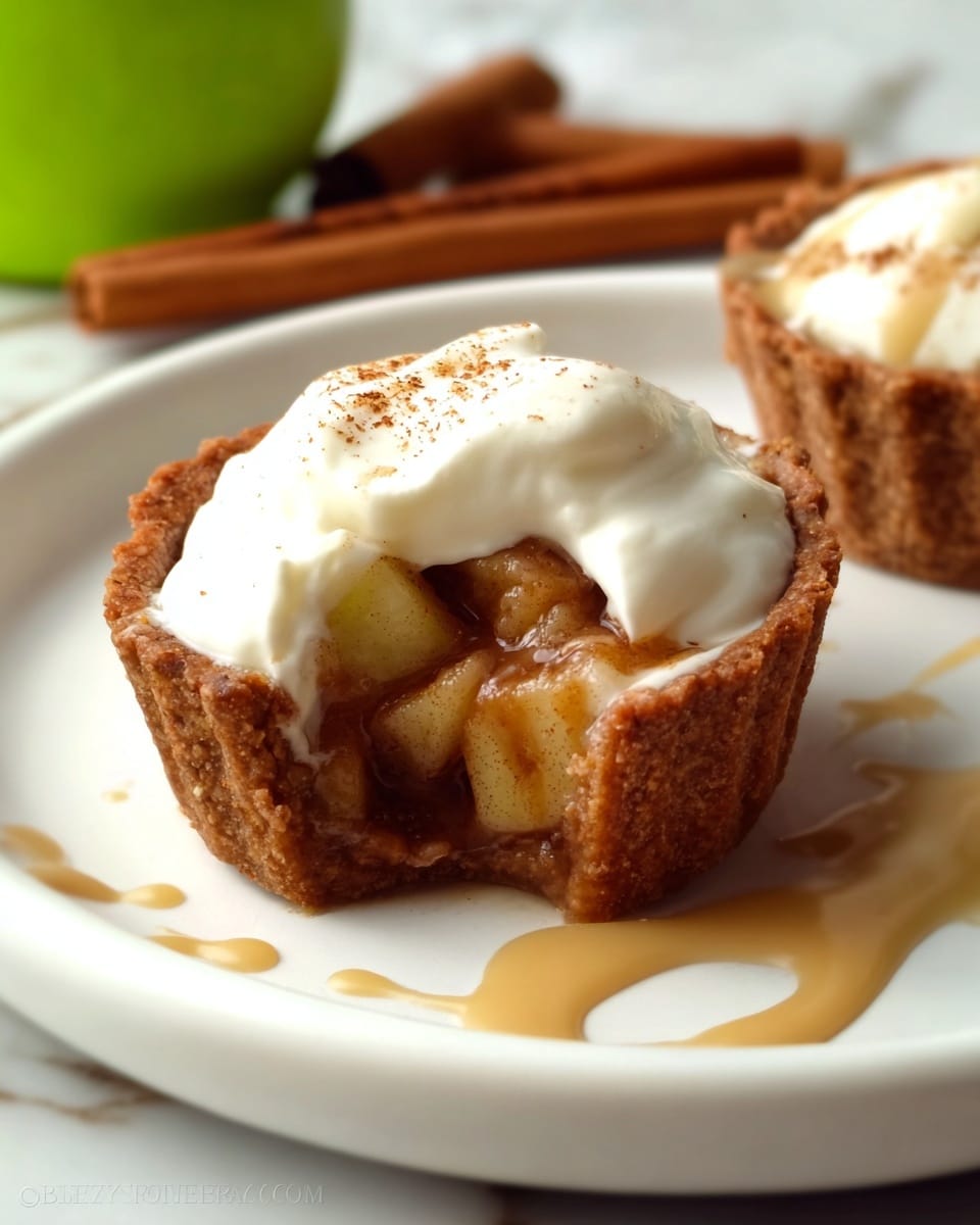 A small tart with a thick, golden-brown crust forms the base, filled with a chunky apple mixture that peeks through the creamy white topping. The topping is soft and smooth, likely whipped cream or yogurt, draped unevenly over the filling and drizzled with a light brown sauce that spills slightly onto the white plate. The plate sits on a white marbled surface with a cinnamon stick and green apple blurred in the background, adding warm and fresh tones to the scene. Photo taken with an iphone --ar 4:5 --v 7