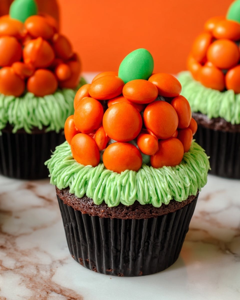 A chocolate cupcake with a black liner forms the base, topped with bright green frosting that looks like grass, made from thin strands piped closely together. On top of the green frosting sits an orange pumpkin-shaped decoration made from round orange candies clustered together, with a small green candy piece on top acting as the pumpkin stem. The cupcakes sit on a white marbled surface with an orange backdrop visible in the background. photo taken with an iphone --ar 4:5 --v 7