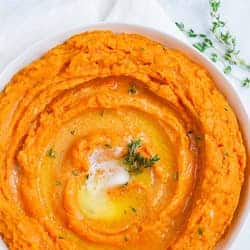 The image shows a glass casserole dish filled with two layers: the bottom layer is bright orange mashed sweet potatoes with a soft texture, and the top layer is a golden brown crumb topping that looks crispy and slightly crunchy. A spoon rests inside the dish, scooping out part of the soft orange layer, revealing the thick and smooth texture beneath the crumb crust. The dish is placed on a white marbled surface with a striped cloth beside it. photo taken with an iphone --ar 4:5 --v 7