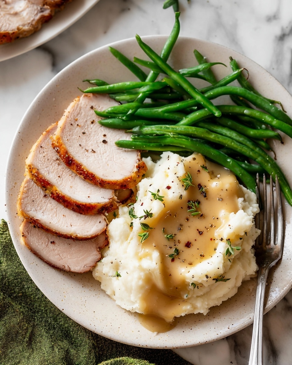 A white plate holds three main sections: at the bottom left, four slices of light brown roasted meat with a slightly darker crust, arranged overlapping; in the center, a mound of creamy white mashed potatoes topped with smooth, light brown gravy and sprinkled with black pepper and small green herbs; at the top, a neat pile of fresh, bright green whole green beans. A silver fork rests on the right side of the plate, touching the mashed potatoes. The plate sits on a white marbled textured surface with a green cloth partially visible below. Photo taken with an iphone --ar 4:5 --v 7
