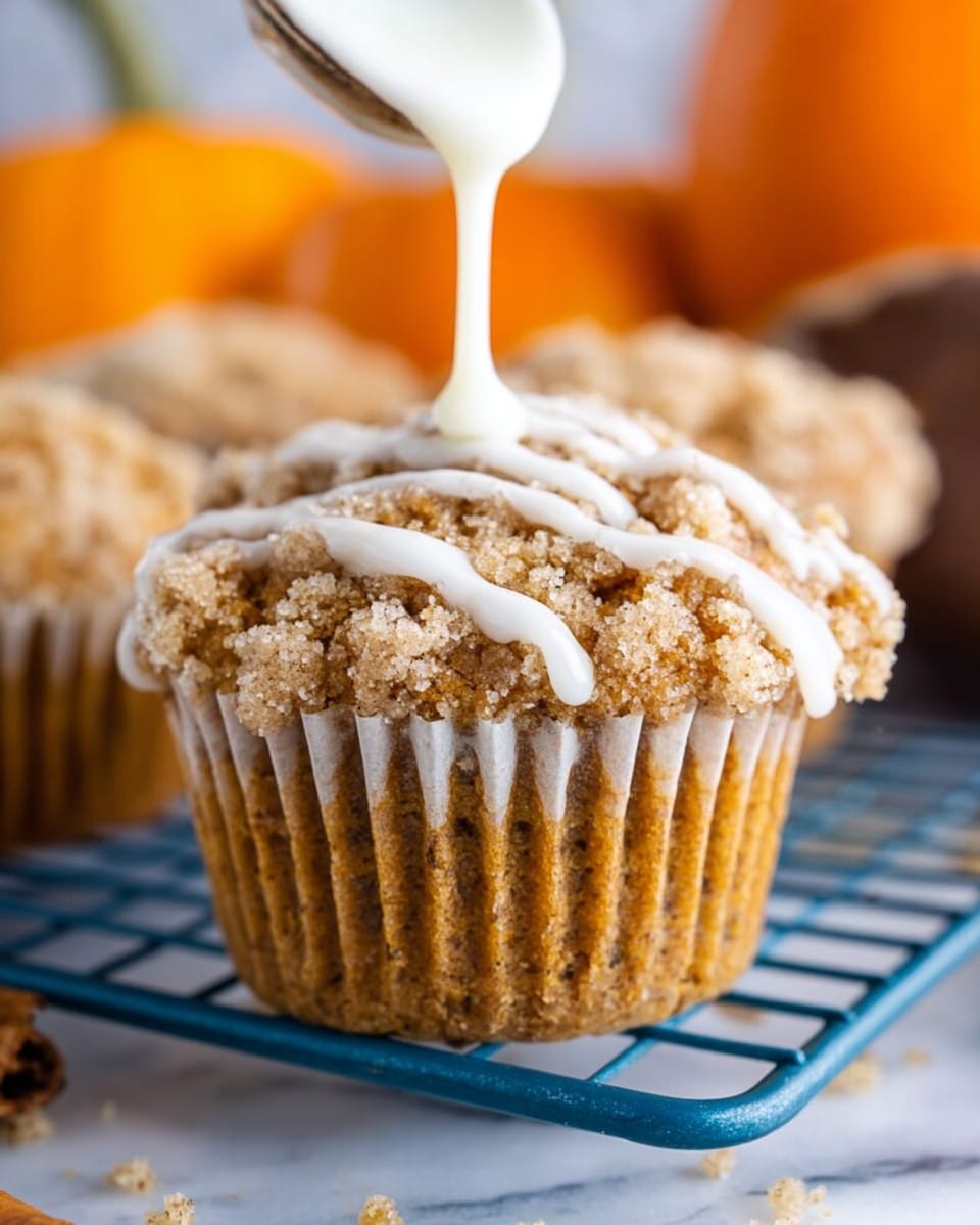 The close-up image shows a single muffin with a crumbly, golden brown topping that looks soft and slightly grainy. White icing is being poured from a white spoon, creating thin, irregular stripes over the crumb layer. The muffin is in a white paper cup with ridges, sitting on a blue cooling rack. The background features a soft focus of orange pumpkins and a brown object, all placed on a white marbled surface. photo taken with an iphone --ar 4:5 --v 7