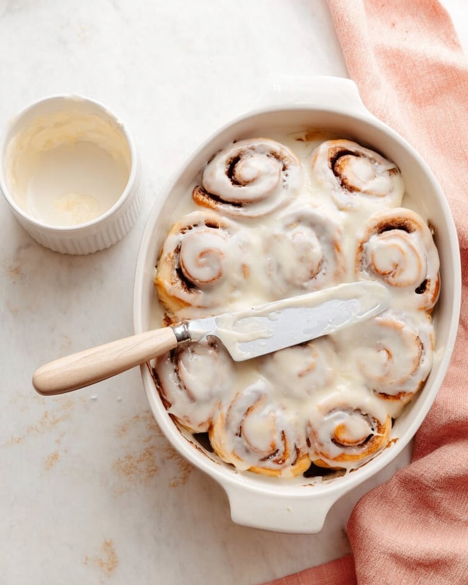 The image shows a white oval baking dish filled with cinnamon rolls covered in creamy white icing. The cinnamon rolls have a golden-brown swirl pattern visible beneath the smooth, shiny icing that spreads over the top layer in soft waves. A silver butter knife with a light wooden handle rests diagonally on the dish, partly on the cinnamon rolls. Next to the dish is a small empty white bowl. The background is a white marbled texture with a light pink cloth partially visible on the right side. Photo taken with an iphone --ar 4:5 --v 7