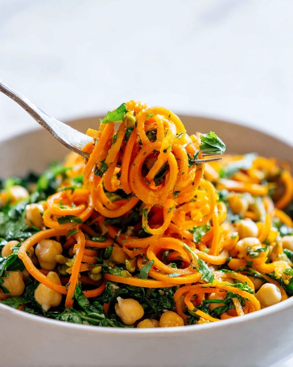 A close-up of a white bowl filled with bright orange spiralized noodles mixed with green leafy vegetables and white chickpeas. There are finely chopped herbs scattered throughout, adding freshness and color contrast to the dish. A fork lifts some of the noodles and greens, showing the curly texture of the noodles and small pieces of green leaves clinging to them. The scene is set on a white marbled surface with soft natural light highlighting the vibrant colors and fresh textures. photo taken with an iphone --ar 4:5 --v 7