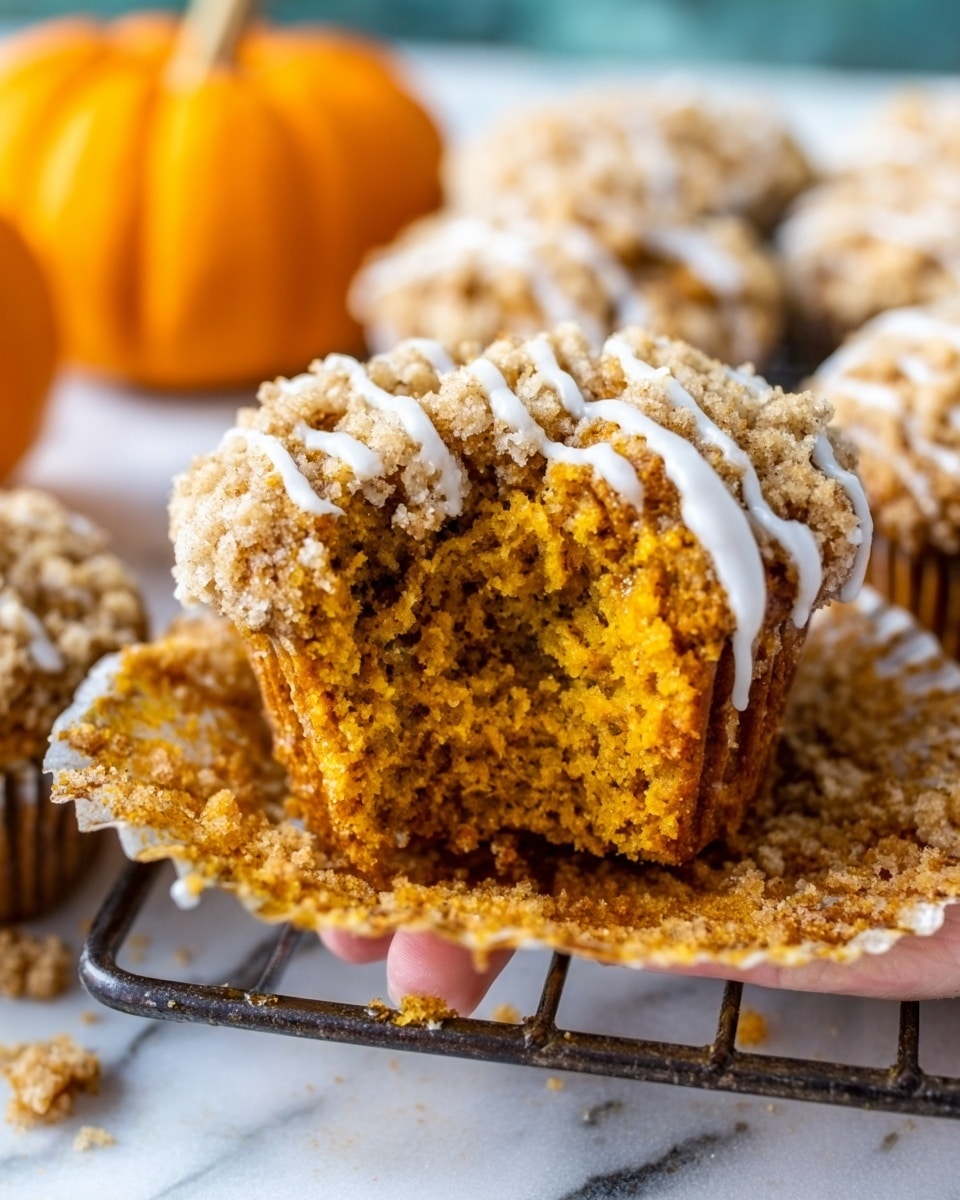The image shows a close-up of a pumpkin muffin with a crumbly, light brown topping that looks soft and slightly crunchy. The inside of the muffin is a warm orange color, moist and fluffy in texture. White icing is drizzled over the top of the muffin in thin lines, adding a sweet contrast to the crumbly layer. The muffin is sitting on a silver wire rack placed on a white marbled surface, with a small orange pumpkin blurred in the background. In the foreground, a woman's hand holds the muffin, showing the open center where a bite has been taken. Photo taken with an iphone --ar 4:5 --v 7