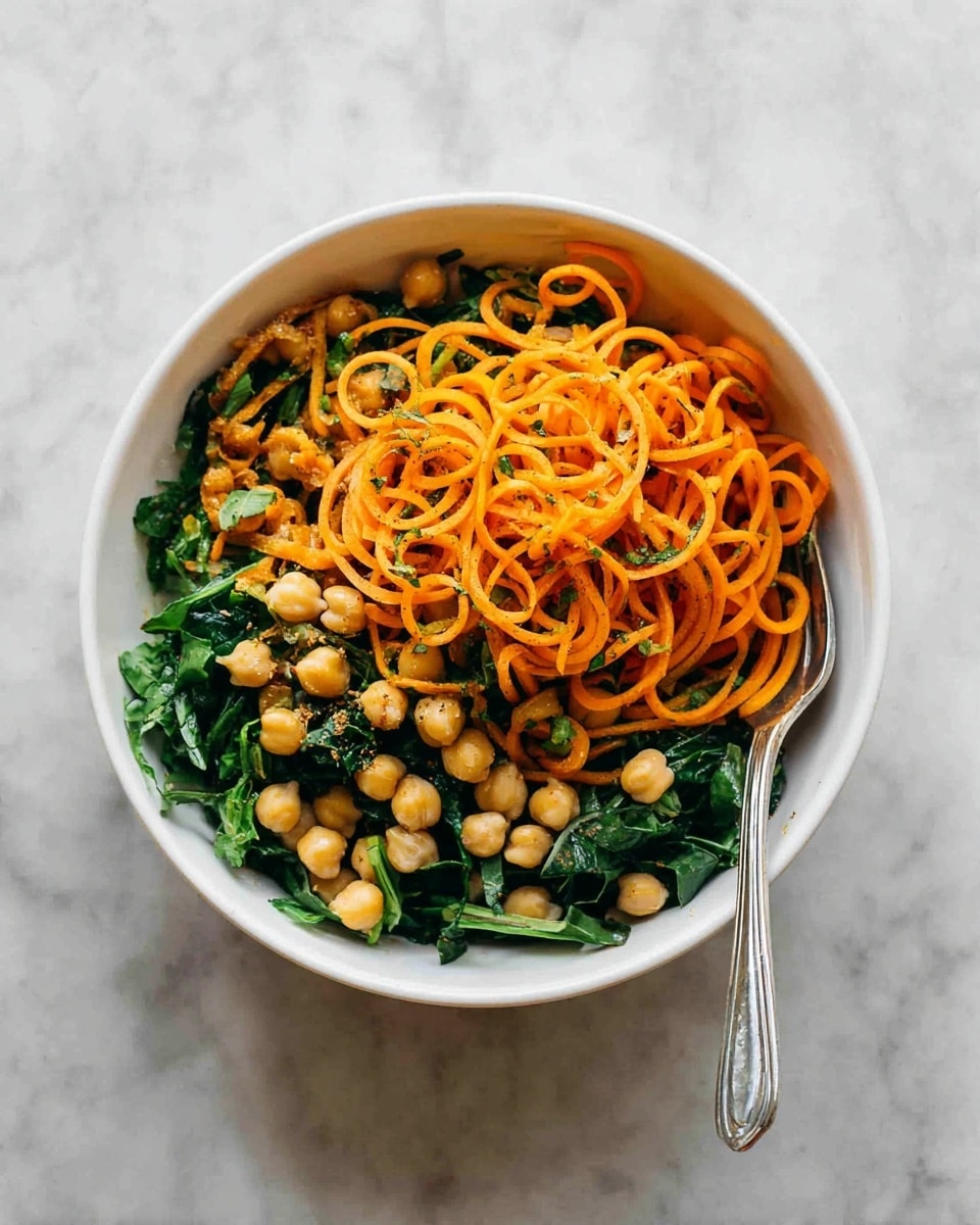 A white bowl filled with three layers of food is placed on a white marbled surface. The bottom layer is chopped dark green leafy vegetables, the middle layer consists of bright orange spiral-shaped vegetable noodles, and the top layer is scattered with round, light beige chickpeas. A silver spoon is resting inside the bowl on the right side. The photo taken with an iphone --ar 4:5 --v 7