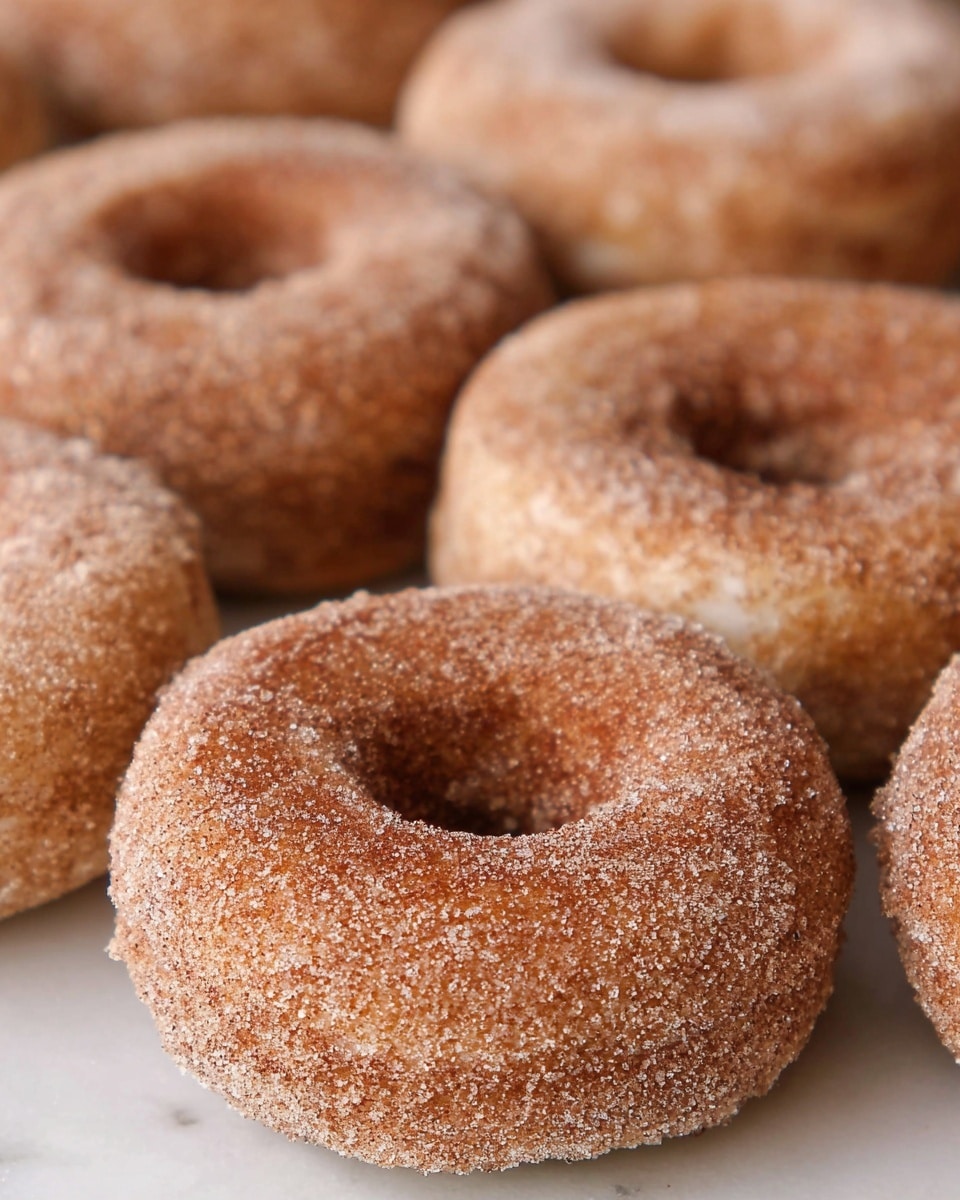 A stack of seven cinnamon sugared donuts is placed slightly off center on a red and white checkered cloth inside a light wooden tray. The donuts are golden brown with a rough sugar coating, giving them a grainy look. Around the stack, more donuts lie on the tray, showing their round shape and hole in the middle. On the left side of the tray, a fresh yellowish-red apple adds color contrast. The background is softly blurred with warm tones, and the surface beneath the tray shows a white marbled texture. photo taken with an iphone --ar 4:5 --v 7